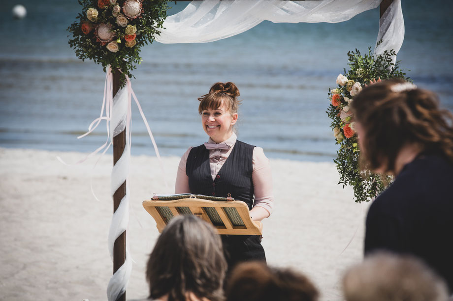 Braut am Strand hält ihre Blumenkinder an den Händen, das Klein weht im Wind, hinten im Bild die Ostsee