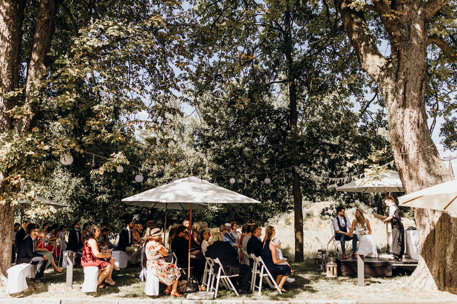 Die Hochzeitsgesellschaft sitzt auf Stühlen. Weiße Sonnenschirme und Bäume spenden Schatten