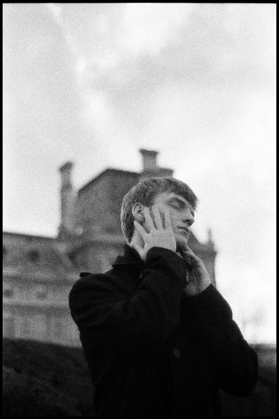 Black-and-white analog fine art portrait of a professional male model with short textured hair, hand resting against his cheek, wearing a dark high-collar jacket.
