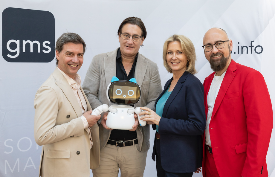 Vier Personen stehen vor einer GMS-Wand und halten gemeinsam den Roboter YUNA.