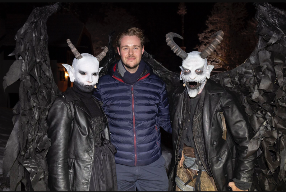 Ein Mann steht zwischen zwei aufwendig als dämonische Krampusfiguren verkleideten Personen mit Hörnern, weiß geschminkten Gesichtern und schwarzen Flügeln.