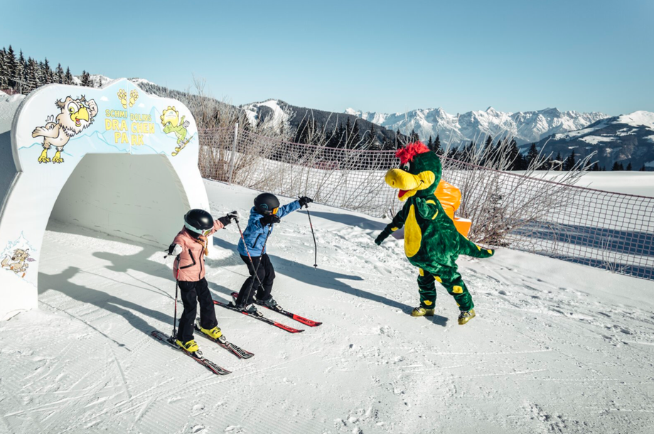 Zwei Kinder fahren durch einen Torbogen in Schmidolins Drachenpark und werden vom Drachenmaskottchen begrüßt.