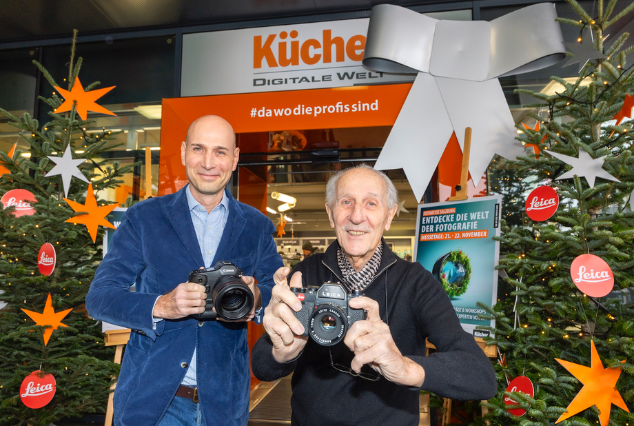 Ulrich Kücher und Franz Kücher halten Kameras vor dem Eingang der Kücher Digitalen Welt in Salzburg, umgeben von weihnachtlicher Dekoration.
