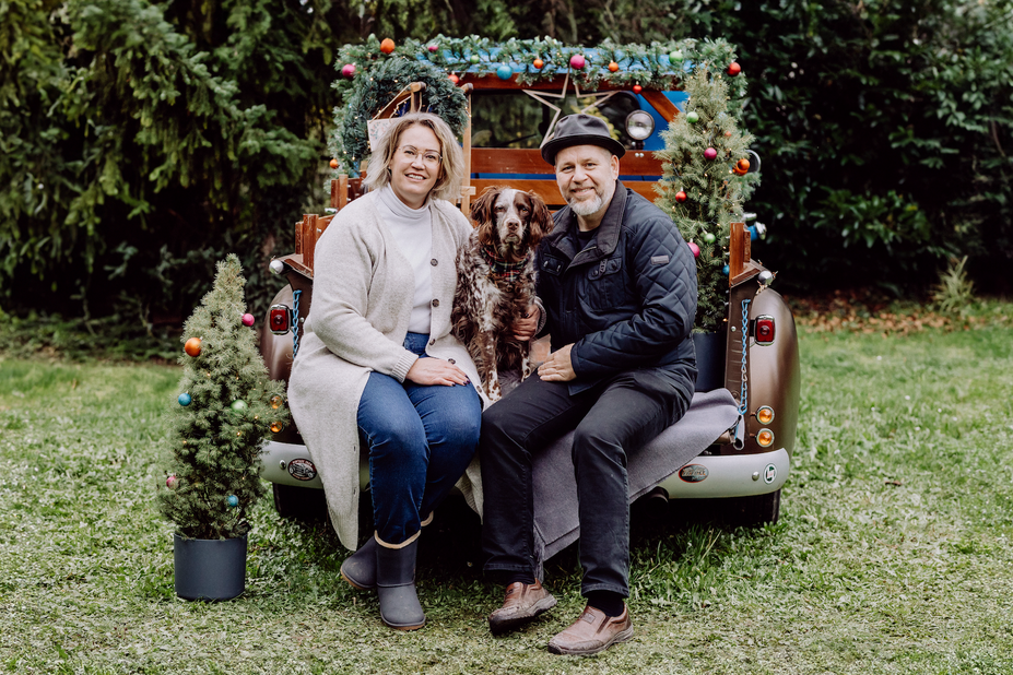Ein Mann mit Hut und eine Frau mit hellen Haaren sitzen auf der Ladefläche eines Pick-Ups, zwischen ihnen sitzt ein Hund, alle schauen in die Kamera. Das Auto ist bunt und weihnachtlich geschmückt.