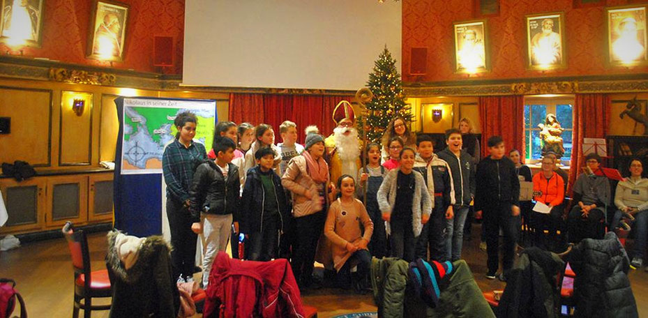 Mädchen und Jungen stehen neben dem Nikolaus und singen gemeinsam ein Lied, rechts im Hintergrund sitzen die Musikanten