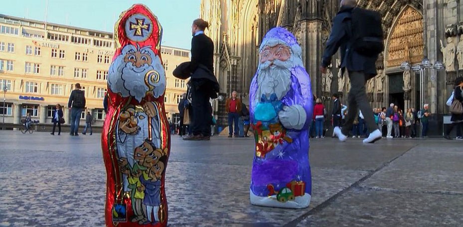 Im Vordergrund steht der rotgekleidete (Schoko)Nikolaus und rechts daneben der lilafarbene (Schoko)Weihnachtsmann. Im Hintergrund sieht man den Kölner Dom, an dem Leute vorbeigehen.