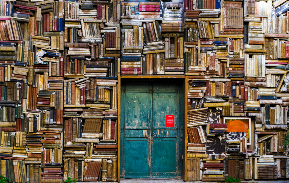 Türkise, alte Tür mit rotem Plakat, komplett umrahmt von einem Bücherregal mit übereinander liegenden alten Büchern und Magazinen