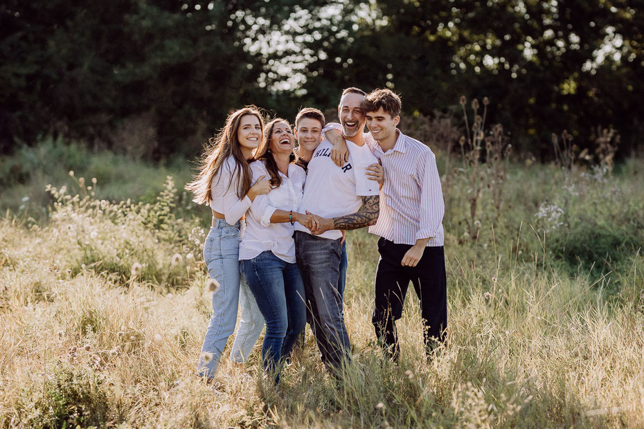 Eine Familie mit drei erwachsenen Kindern steht auf einer Wiese im Sommerlicht, dabei umarmen sie sich und lachen fröhlich