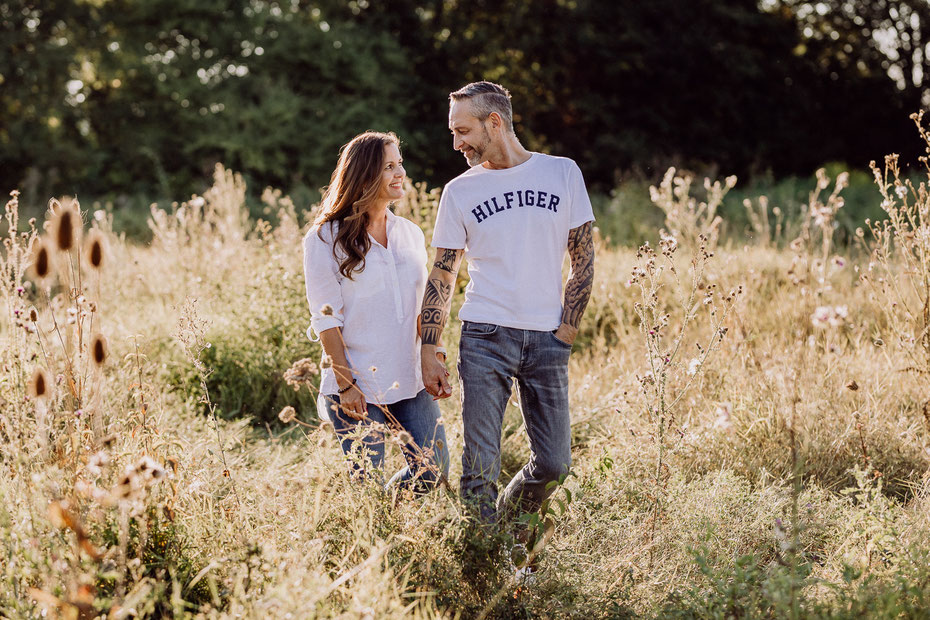 Vater und Mutter stehen auf einem Feld bei Familienshoot in Rheinhessen, die Sonne steht tief, beide sind mit blauer Jeans und weißem Oberteil gekleidet, sie halten sich an der Hand und schauen sich lächelnd an.