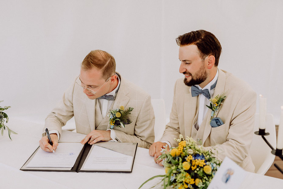 zwei Bräutigame in beigefarbenen Anzügen sitzt an einem Tisch, der links sitzende Bräutigam unterschreibt die Eheurkunde, der rechts sitzende Bräutigam schaut ihm dabei zu