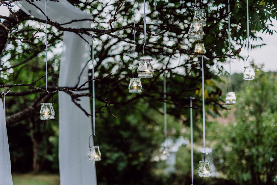 Nahaufnahme von Kerzenhaltern aus Glas, in denen kleine Teelichter brennen und die an weißen Bändern befestigt in eine Baumkrone gehängt wurden