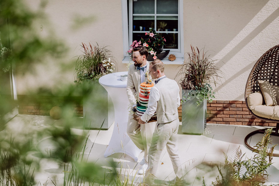 zwei Bräutigame in beigefarbenen Anzügen tragen gemeinsam eine Hochzeitstorte in Regenbogenfarben über eine Terasse