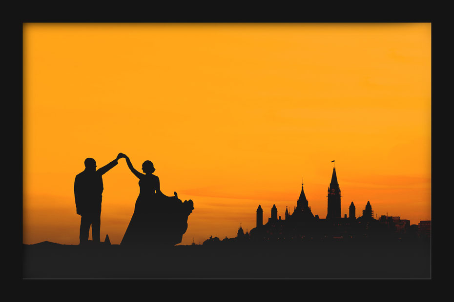 Wedding couple in silhouette dancing in front of Parliament hill in Ottawa. She is wearing a dress he is wearing a suit. Sky is orange. 