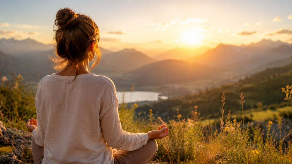 Frau sitzt meditierend auf einem Hügel mit Blick auf Berge und einen See im warmen Abendlicht – Symbol für innere Ruhe, Selbstheilung, seelische Gesundheit, Achtsamkeit und den Weg zurück zu sich selbst.