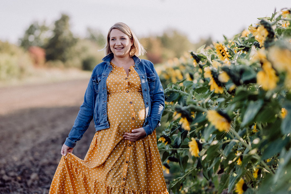 Junge, schwangere Frau geht entlang eines Sonnenblumenfelds auf die Kamera zu 