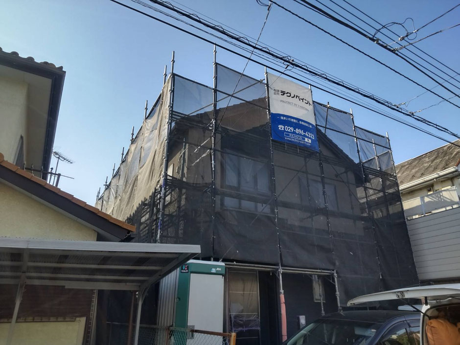 外壁塗装中家、足場と黒い養生ネットが設置され、「テクノペイント」の現場シートが設置されている