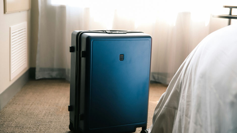 Ein Koffer steht neben einem Bett. Bild von Taylor Beach auf Unsplash.