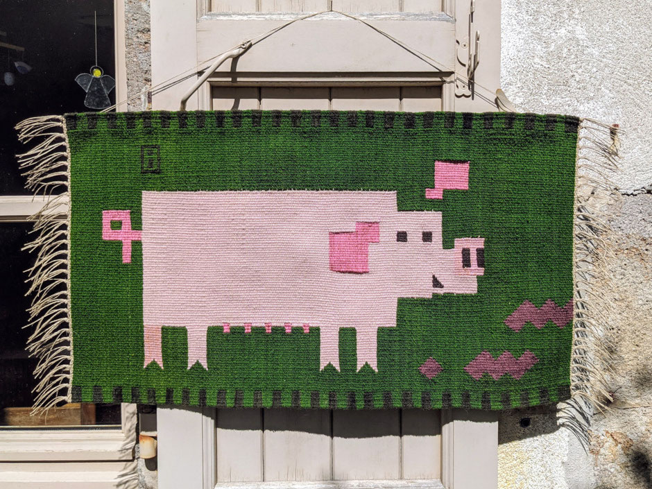 Un kilim-tenture murale en tissage, au dessin géométrisé,  au fond vert et une mère cochon rose. Tissage mural accroché sur un volet en bois à l'extérieur.