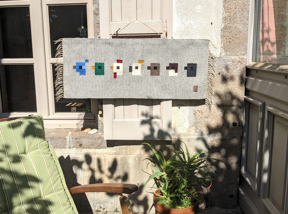 Sur une terrasse le kilim-tenture murale en tissage représentant une vache salers qui regarde un papillon sur un pré de crocus est accroché sur un volet. Dessin du tissage mural géométrisé. Un aloès et une chaise-longue sont visibles devant.