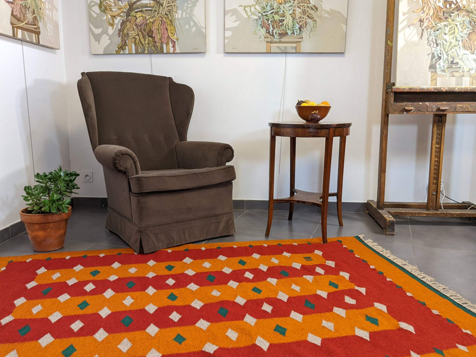 Intérieur avec, sur le sol, un kilim rouge-orange, motif diamants gris et vert. À droite, une table avec un bol et les fruits, au-dessus, trois tableaux - nature morte, à gauche une plante succulente en pot et un fauteuil voltaire au centre.