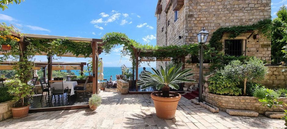 An outdoor seating area with wooden tables and chairs under a pergola covered with green vines. Beautiful sea view of the sea, with a backdrop of blue sky, scattered clouds, stone buildings, and decorative plants in pots, creating an inviting atmosphere