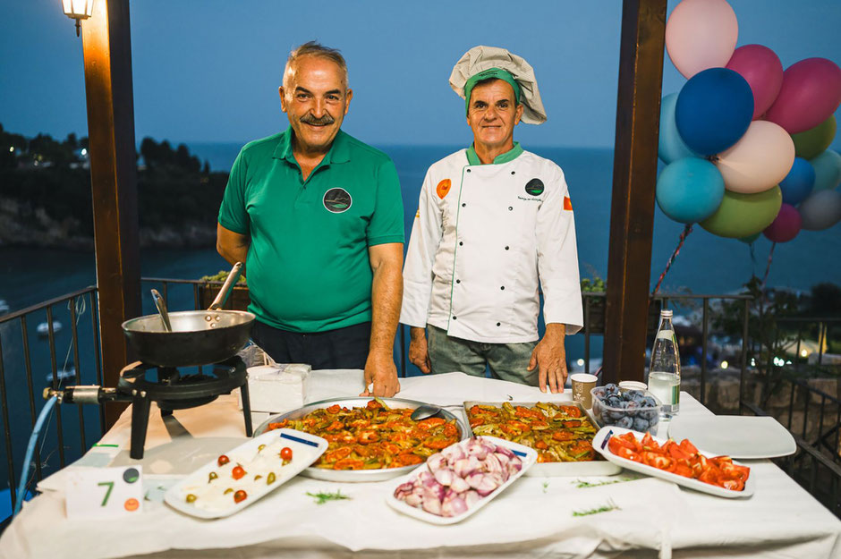 Two chefs standing behind a table with traditional okra dishes, fresh vegetables, and cooking utensils, with the sea and balloons in the background during Okra Days at Palata Venaziain Hotel in Ulcinj