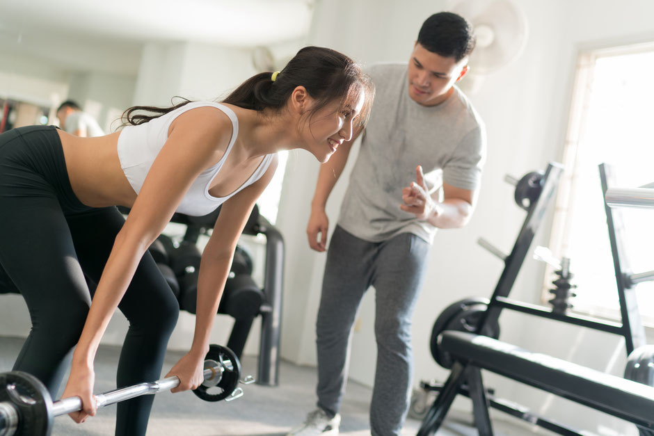 A young asian girl practicing her dead lift using a barbell while her male personal trainer monitors if she is performing the movement correctly