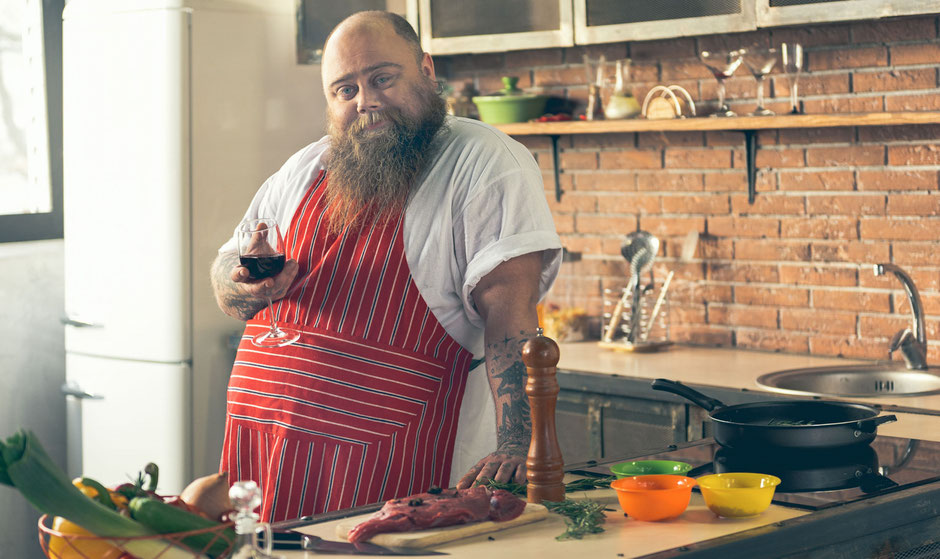 Gicht-Ursachen: Dicker Mann in Küche mit Weinglas und Fleisch