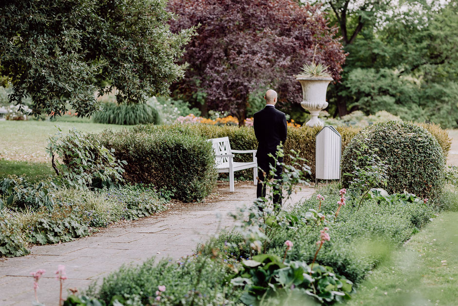 Bild eines Bräutigams, der in einem mit Bäumen und Sträuchern bewachsenen Park mit dem Rücken zum Betrachter steht und wartet.