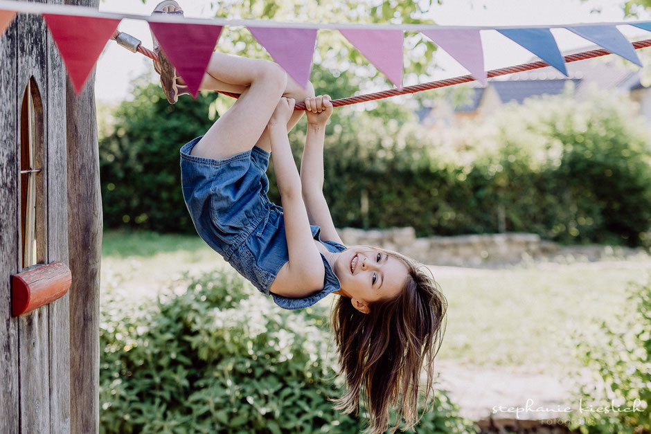 ein braunhaariges Mädchen in einem Jeanskleid hängt mit Händen und Beinen an einem Seil auf einem Spielplatz 