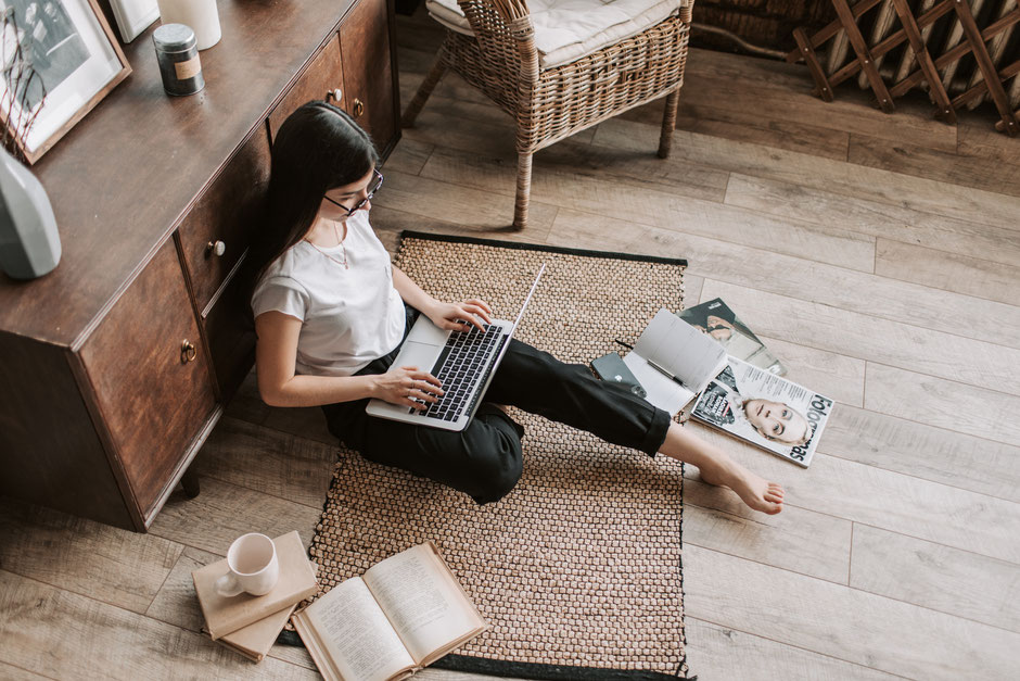 Vrouwelijke copywriter zit op de grond met rug tegen de kast en haar laptop op schoot en schrijft webteksten.