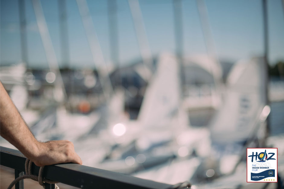 Hand eines HOZ Inside Members am Geländer mit Blick auf Yachthafen – Segelboote im Hintergrund, maritime Atmosphäre am Steg