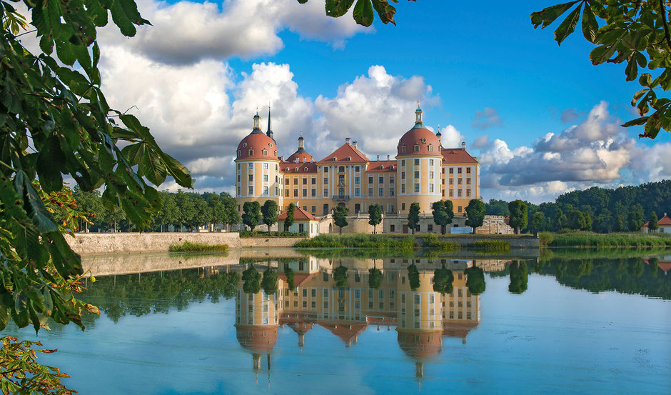 A water castle photographed in beautiful weather. In the blue sky are many white and gray clouds. There are trees everywhere in the picture, there is a lake or pond in front.