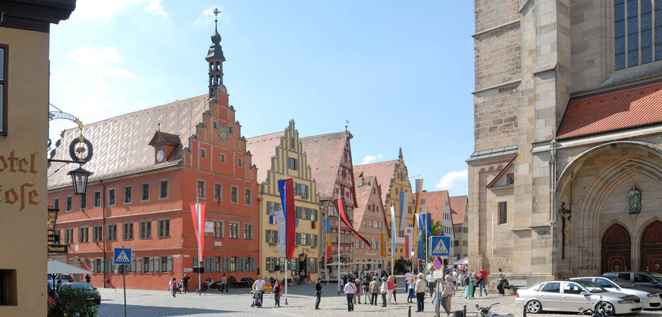 It is the world-famous Dinkelsbühl with its medieval buildings on the way to the historic marketplace. You can see many flags flying and in the foreground some cars are parked and tourists are walking. On the right you can see the part of a church.
