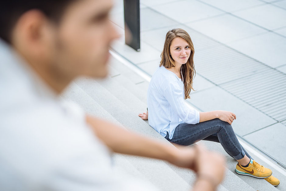 Junge Frau schaut auf einer Treppe liebevoll zu ihrem Partner hinauf, der über ihr sitzt.