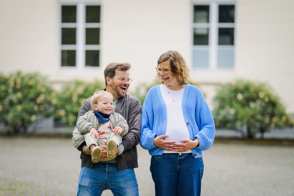Lustiges Babybauch-Shooting: Der Vater imitiert einen Babybauch mit seinem kleinen Sohn neben der schwangeren Mutter.