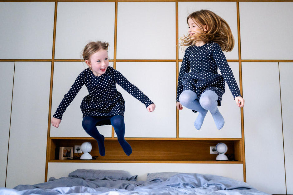 Zwei junge Maedchen springen auf dem Bett der Eltern hoch in die Luft und haben dabei viel Spass.