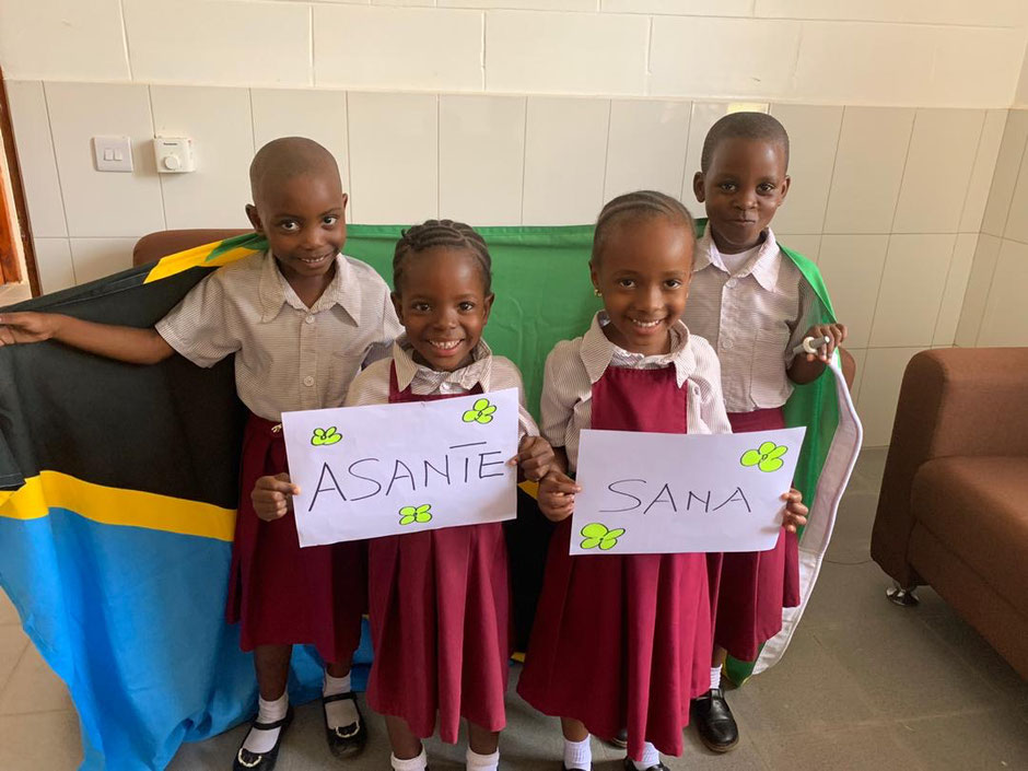 Vier Kinder mit einer Flagge und Schilder mit "Asante Sana" lachen in die Kamera.