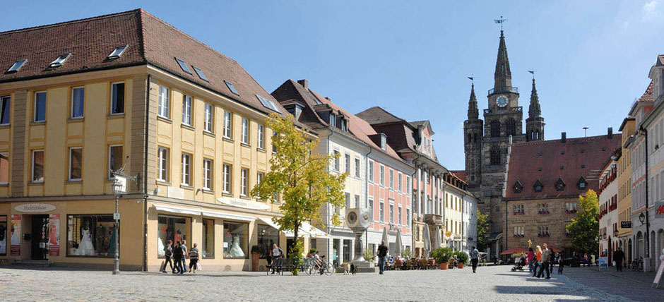 Eine bekannte Ansicht in Ansbach. Eine Fußgängerzone mit hübschen histroischen Gebäuden rechts und links führt auf ein querstehendes Haus, mit Sandsteinen gebaut, zu. Dahinter ragt eine riesige Kirche mit drei Türmen in den Himmel. Man sieht einige Leute.