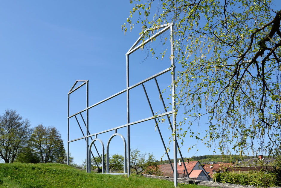 Ein Modell aus Metallstangen steht senkrecht und bildet die Umrisse eines Römer-Kastell-Eingangs mit zwei Türmen. Im Hintergrund sieht man Häuser, Bäume und wolkenlosen, blauen Himmel.