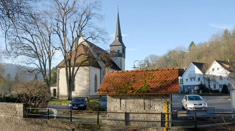 Inmitten von Bäumen ohne Laub steht die Kirche in Widdern. Zwischen ihr und dem Betrachter ist eine halbgeschlossene, historische Bushaltestelle. Rechts sind zwei aktuelle Wohnhäuser, drei Autos parken an der Straße.