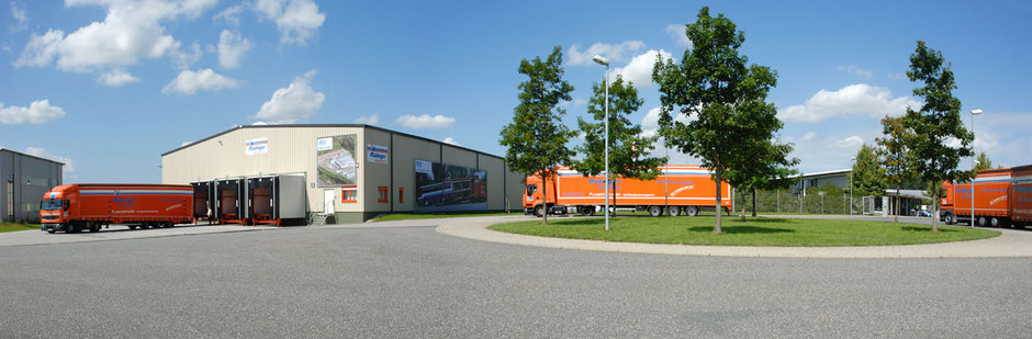Es ist eine riesige runde Grünfläche, um die ein Kreisel geteerte Straße herumführt. Auf der Grünsfläche stehen Bäume und es wächst Gras. Im Hintergrund sieht man drei Lkw mit langen Anhängern. Einer steht vor einer Lagerhalle.