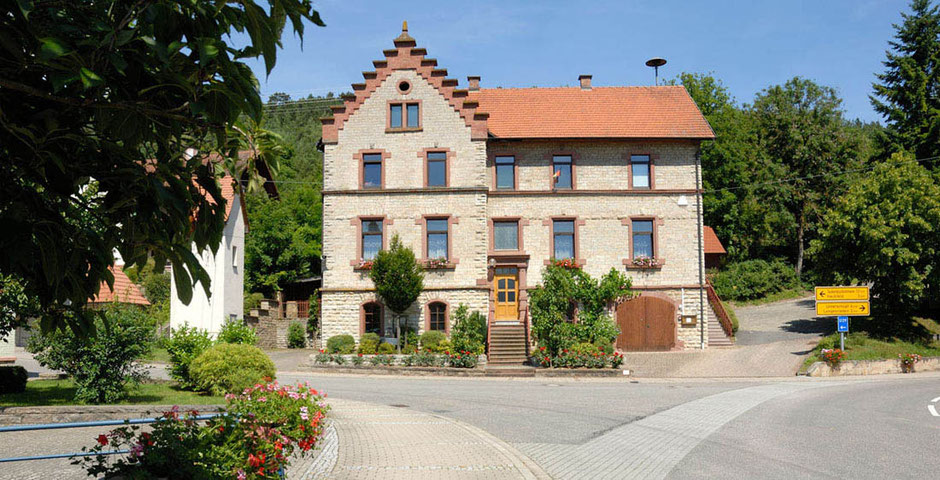 Blick auf ein wunderschönes historisches Gebäude in Kupprichhausen, die Straße gabelt sich davor, rechts sieht man ein Verkehrsschild. Links vorne sind Blumen und ein Baum. Die Sonne scheint, der Himmel ist blau.