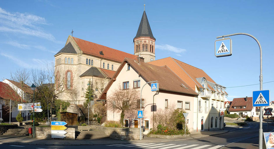 Vielleicht die schönste Ansicht für Fotografen von Assamstadt. Über eine Kreuzung hinweg schaut man auf eine Kirche hinter einer Wohnhauszeile. Im Vordergrund ist ein Zebrastreifen. Man sieht viele Verkehrsschilder. Die Sonne scheint, der Himmel ist blau.
