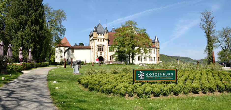 Die Götzenburg in Jagsthausen im Sonnenschein. Um das Schloss sieht man eine Gartenanlage, links sind grüne Bäume und nicht entfaltete Schirme. Der Himmel ist blau mit dünnen Schleierwolken.