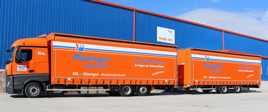 Ein imposanter Lang-Lwk ist von der Seite fotografiert. Natürlich ist er fast komplett orange. Er steht paralell zu einer Neubau-Lagerhalle in der Farbe Blau mit orangenen Anteilen.