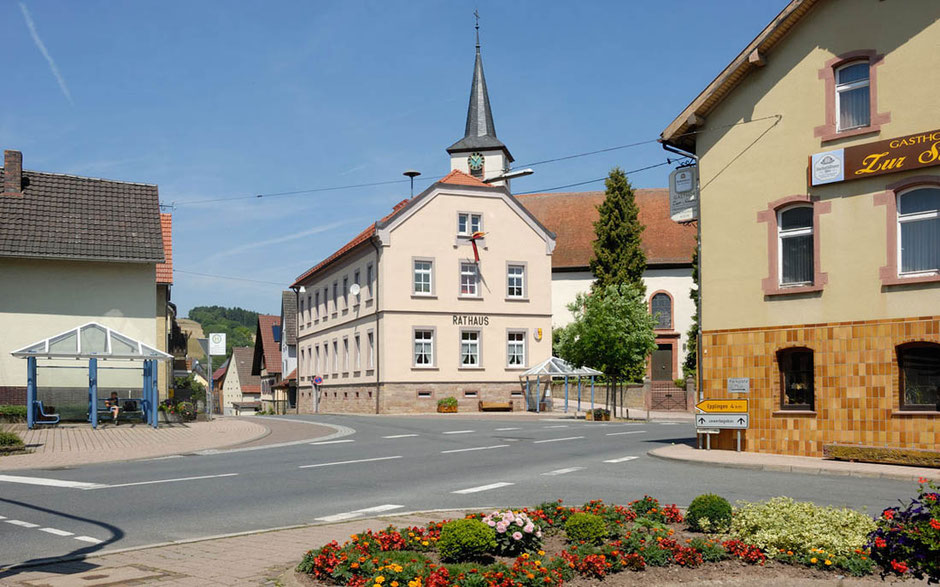 In Schweigern nimmt das halbe Bild vorne die Straße ein, von links nach rechts. Dahinter sieht man das historische Rathaus aus Sandstein. Links ist eine blaue Bushaltestellle, im Vordergurnd ist ein Beet mit vielen Blumen. Im Hintergrund die Kirche.