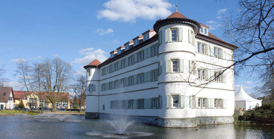 Das Wasserschloss in Bad Rappenau. Man sieht mehrere Springbrunnen, die Bäume sind noch unbelaubt. Eine Seite des Schlosses ist in der Sonne. Rechts ist ein Partyzelt in Weiß aufgebaut. Im Hintergrund sind Häuser.