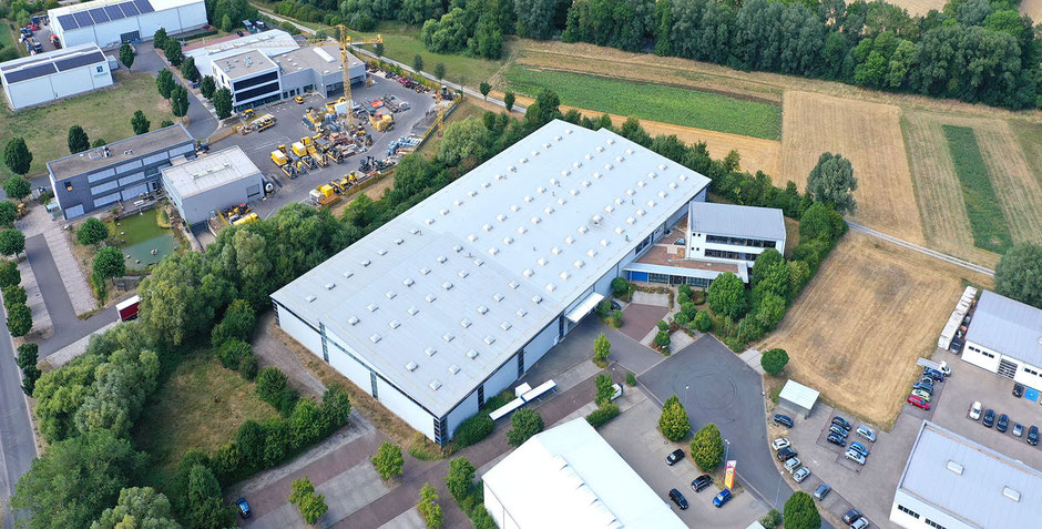 Eine gewaltige Lagerhalle ist in der Bildmitte zu sehen: Es ist ein Foto von einer Drohne fotografiert. Links ist der Komplex eines anderen Eigentümers. Rechts und rechts oben sind Felder.