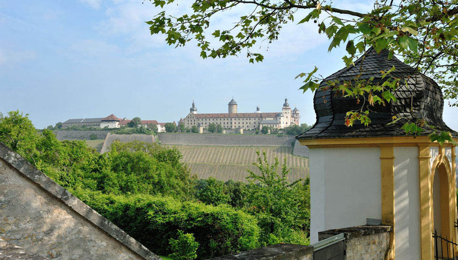 Die Festung Marienberg in Würzubrg. Am Hang davor sieht man einen Weinberg, zwischen dem Weinberg und dem Betrachter ist Wald. Rechts im Wald ist der Turm eines kleinen barocken Gebäudes in Gelb undf Weiß.